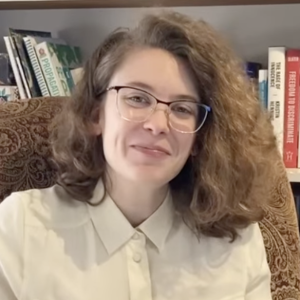 Lee Tremblay, seated in front of bookshelves in an image captured from a video. They have brown wavy hair, glasses and wear a dress shirt.